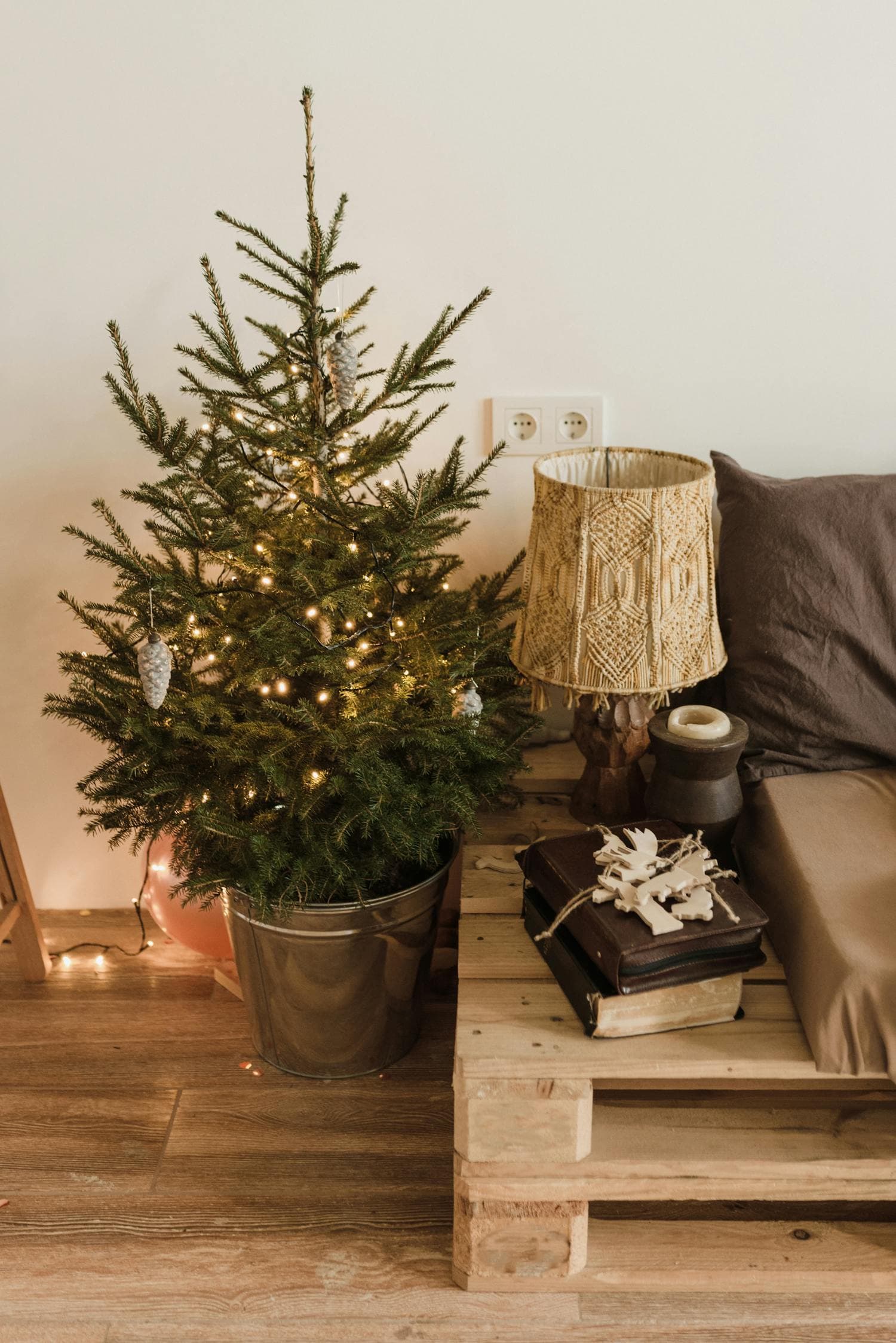 Small live Christmas tree in a galvanized bucket wrapped in warm-white string lights, beside a taupe sofa and a wooden pallet side table with a macrame lamp, a cozy small-apartment Christmas corner
