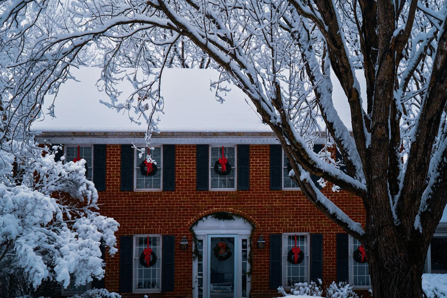 Two-story brick colonial home with symmetric facade, black shutters, snow-covered roof, and a wreath with a red bow hung in every front-facing window