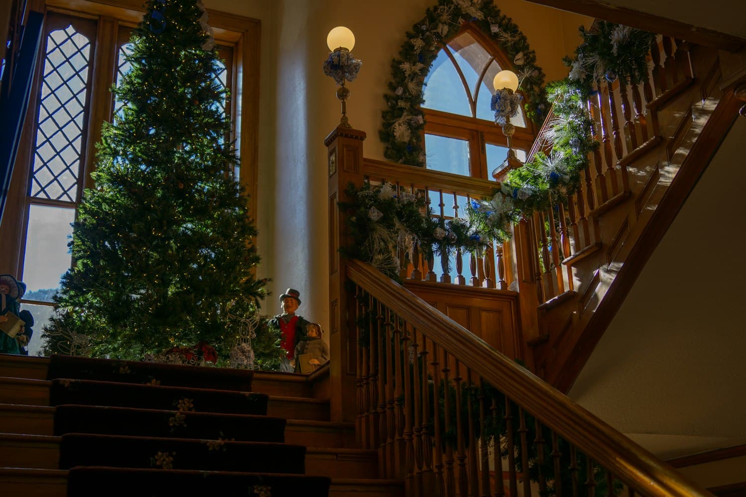 Victorian-style grand staircase with carved wooden banister wrapped in evergreen garland, a tall lit Christmas tree beside a Gothic-arched window, and a Dickensian carol-singer figurine