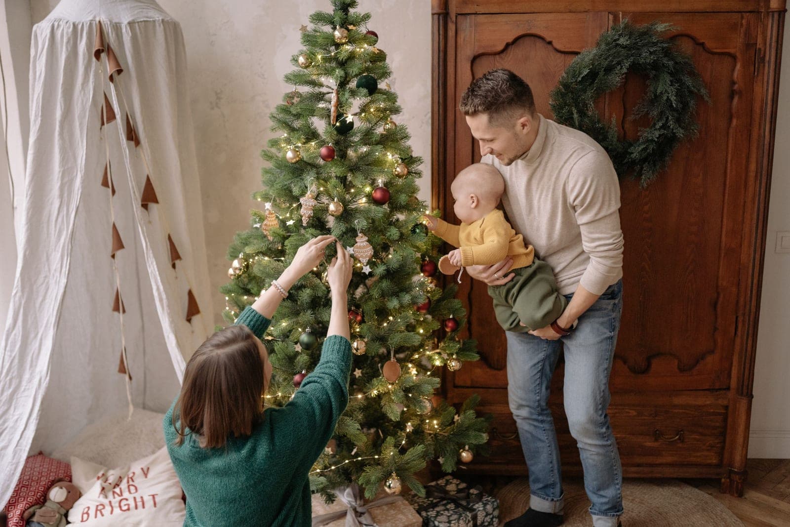 Two parents, one holding a baby, hanging ornaments on a lit Christmas tree beside a dark wood armoire with an evergreen wreath on it