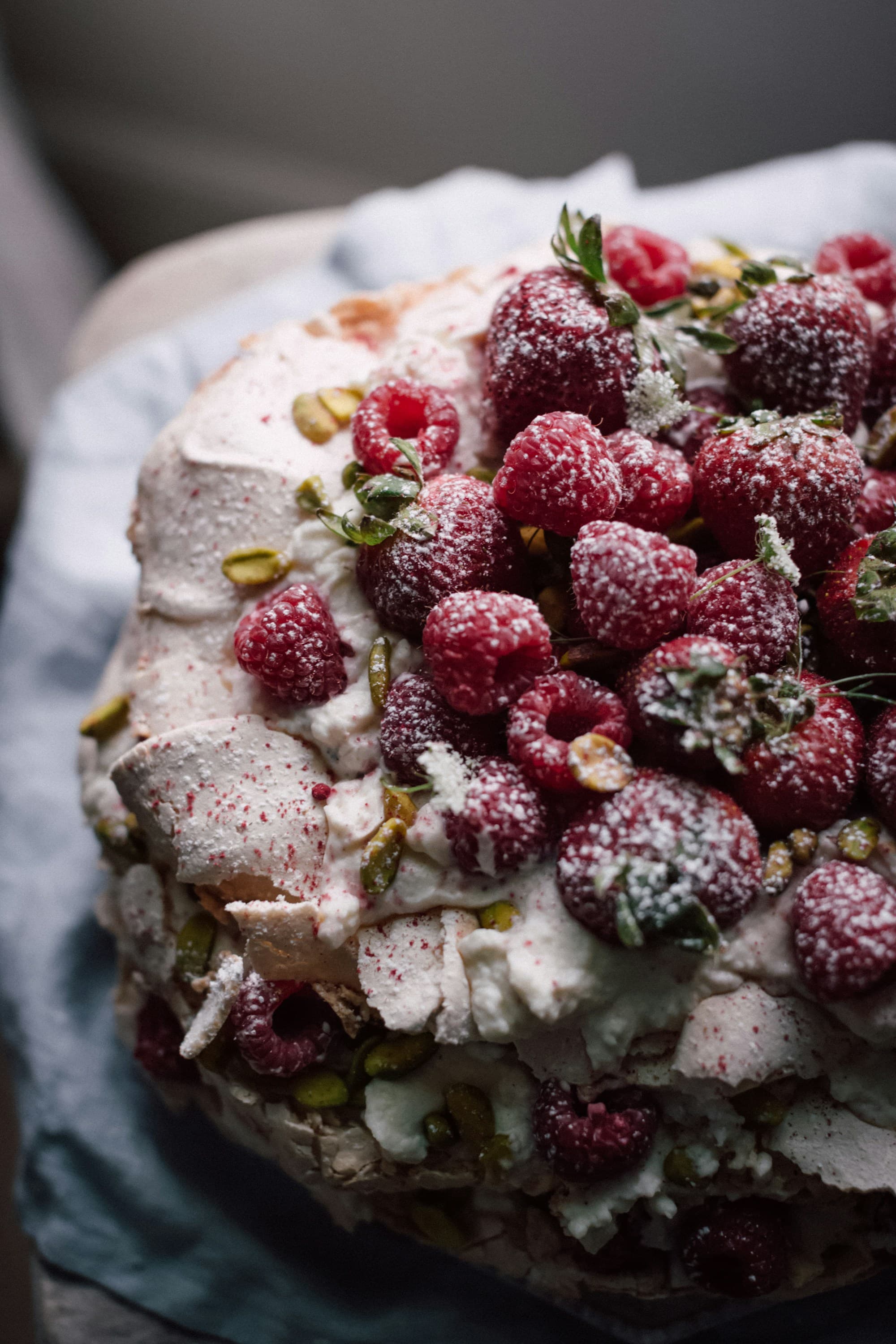 A rustic pavlova piled high with fresh raspberries, scattered pistachios, and torn meringue shards, dusted with powdered sugar and finished with sprigs of green herbs, photographed close on a pale blue linen napkin