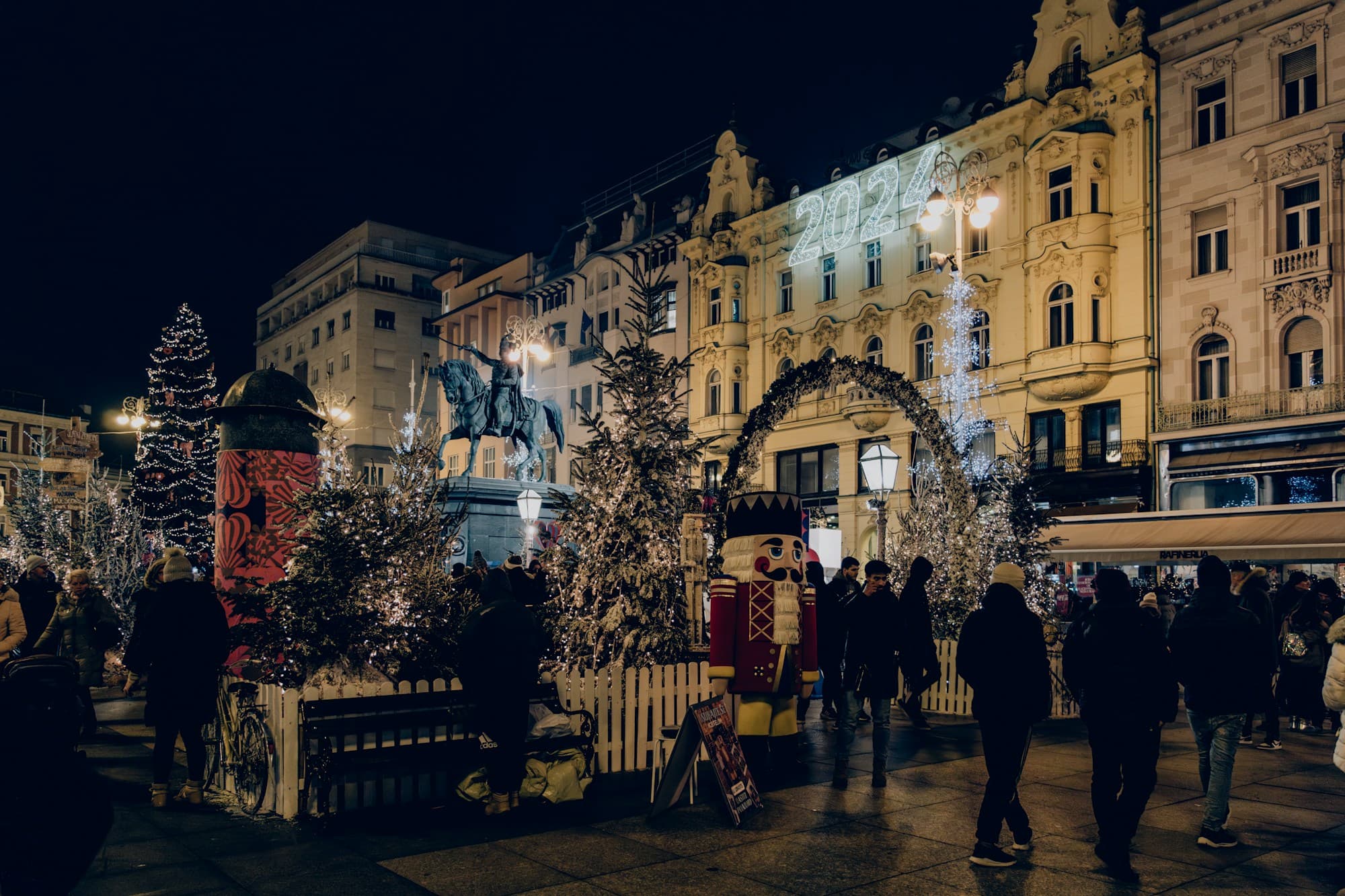 Zagreb's main square during Advent at night, with illuminated Christmas trees, a giant nutcracker, a lit archway, a "2024" light display on a grand belle-epoque building, the equestrian statue of Ban Jelačić, and crowds of visitors among the festive lights, at Zagreb's repeatedly award-winning Christmas market