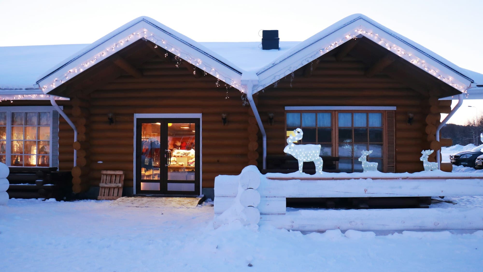 A snow-covered log cabin lit with warm Christmas lights at dusk in Finland