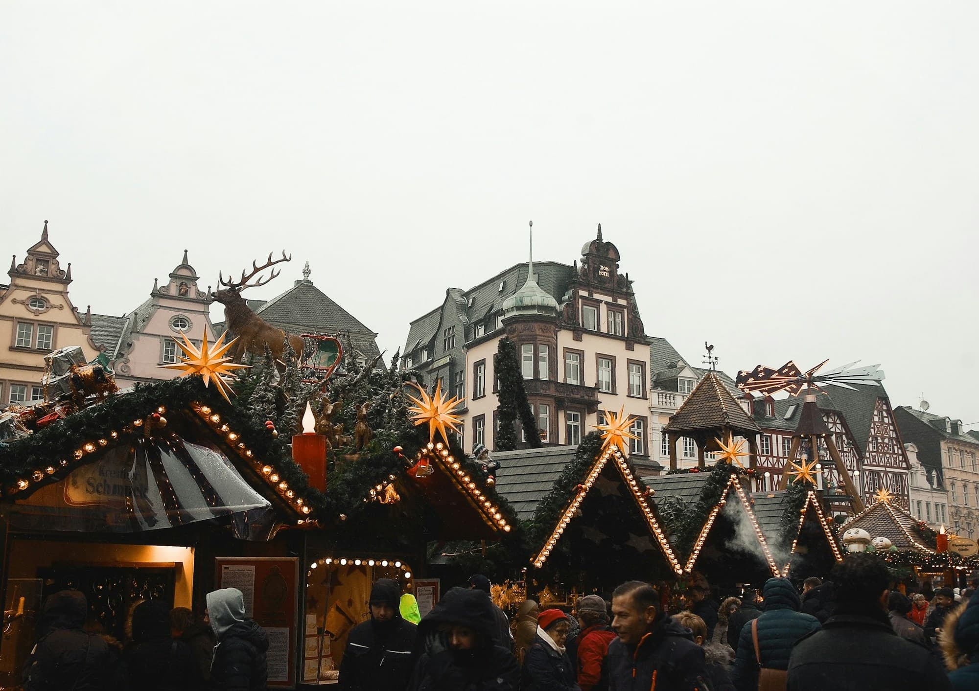 A traditional German Christmas market with rows of wooden gabled stalls strung with white lights and yellow Moravian stars, evergreen garlands draped along the eaves, gabled half-timbered buildings rising in the background, and crowds of bundled visitors browsing