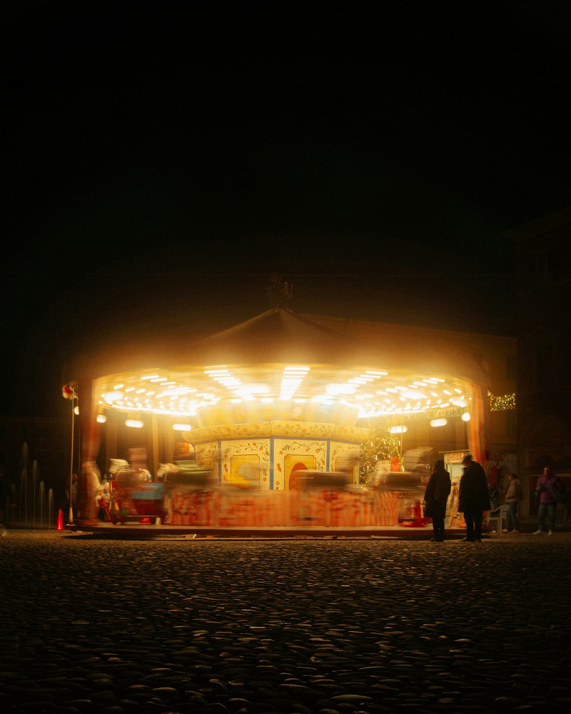 A vintage Italian carousel glowing warm yellow against a dark night sky, set on cobblestones in a European market square, with figures standing in silhouette watching