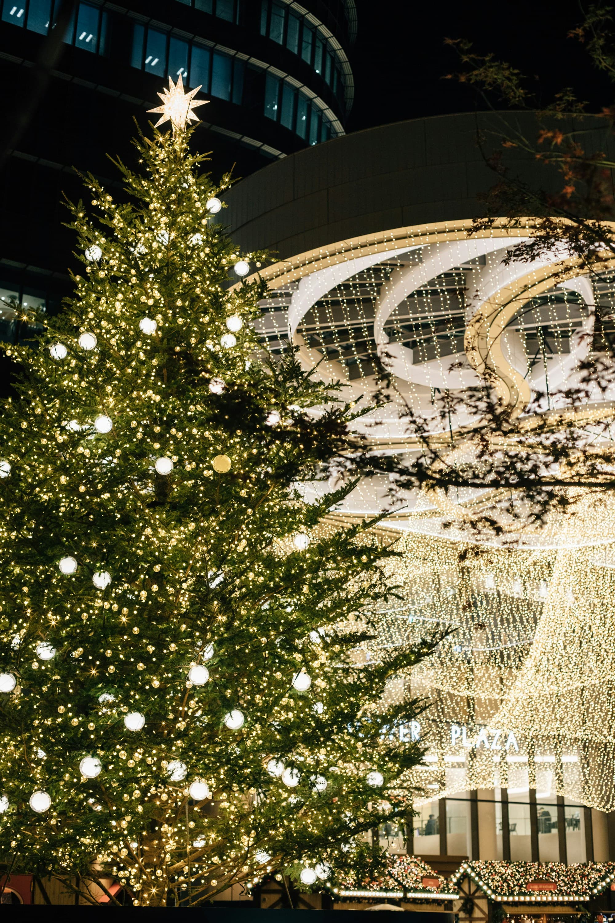 A massive illuminated Christmas tree with a glowing star topper standing in front of a modern Tokyo building, with elaborate white architectural illumination behind it