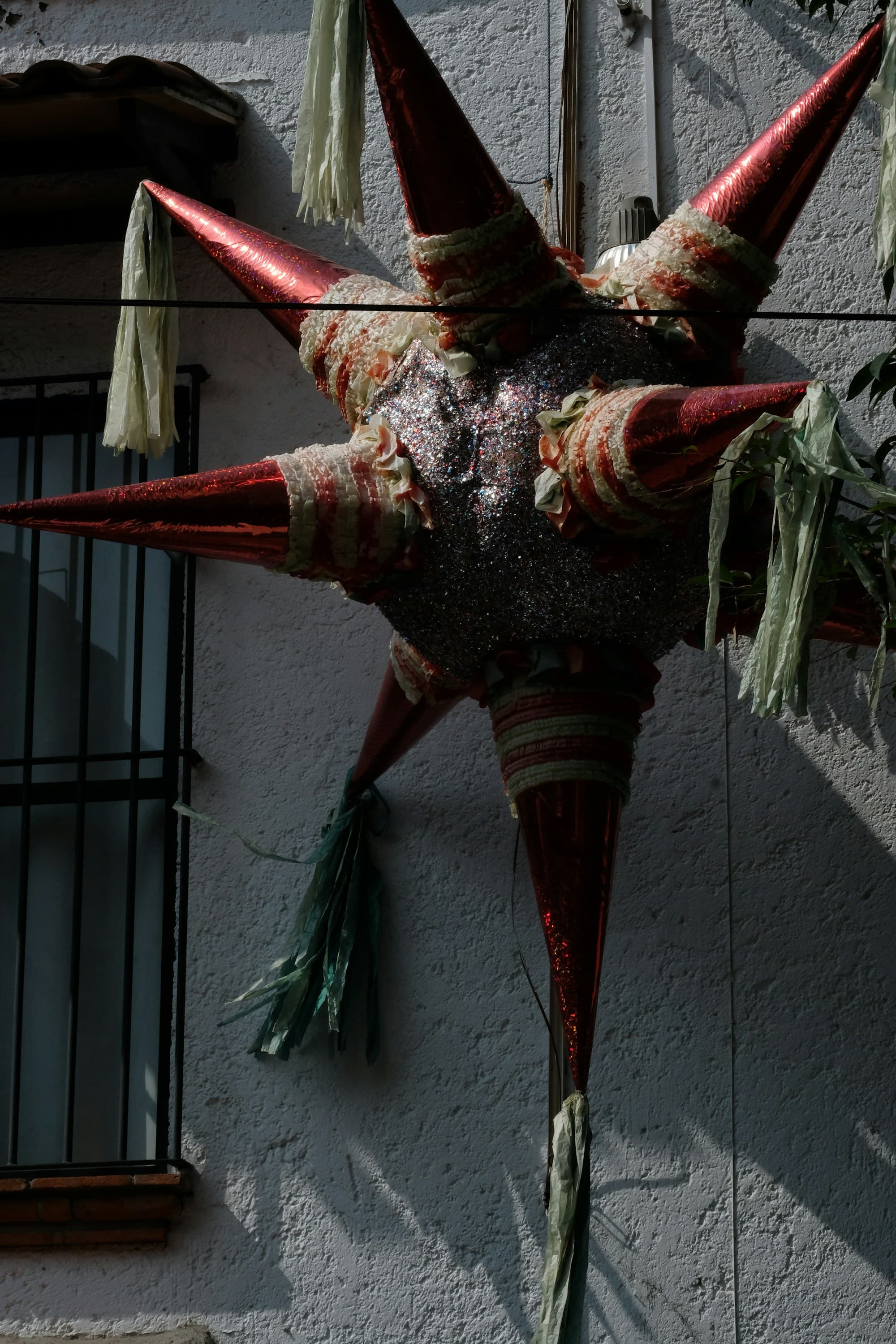 A large traditional Mexican star-shaped piñata in metallic red with green and white paper streamers trailing from each point, hanging against a white stucco wall with a wrought-iron window grille beside it