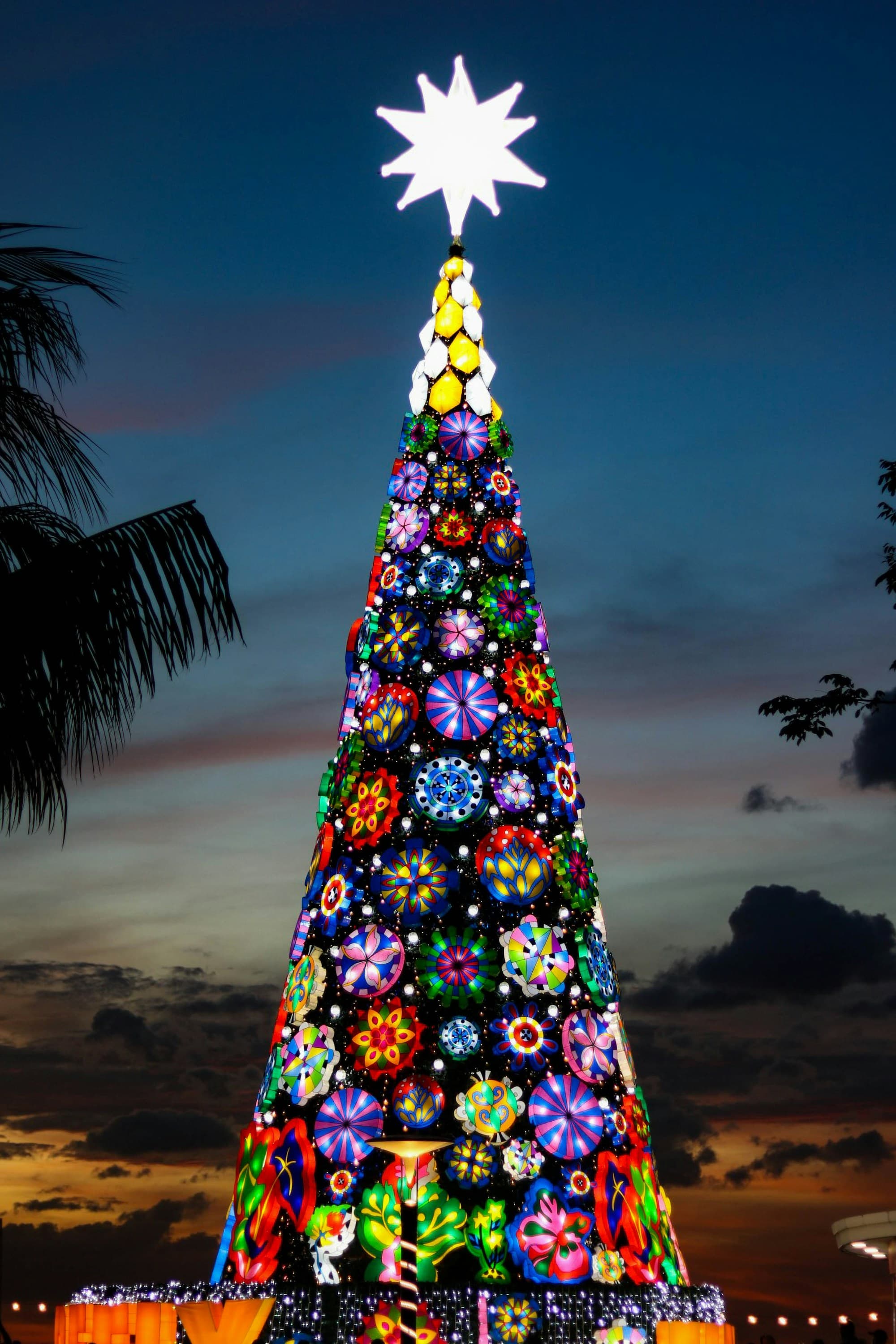 A towering cone-shaped Manila Christmas tree built from dozens of colorful illuminated parol star lanterns in mandala-like patterns, topped by a giant white star, photographed at twilight with palm tree silhouettes against an orange sunset sky
